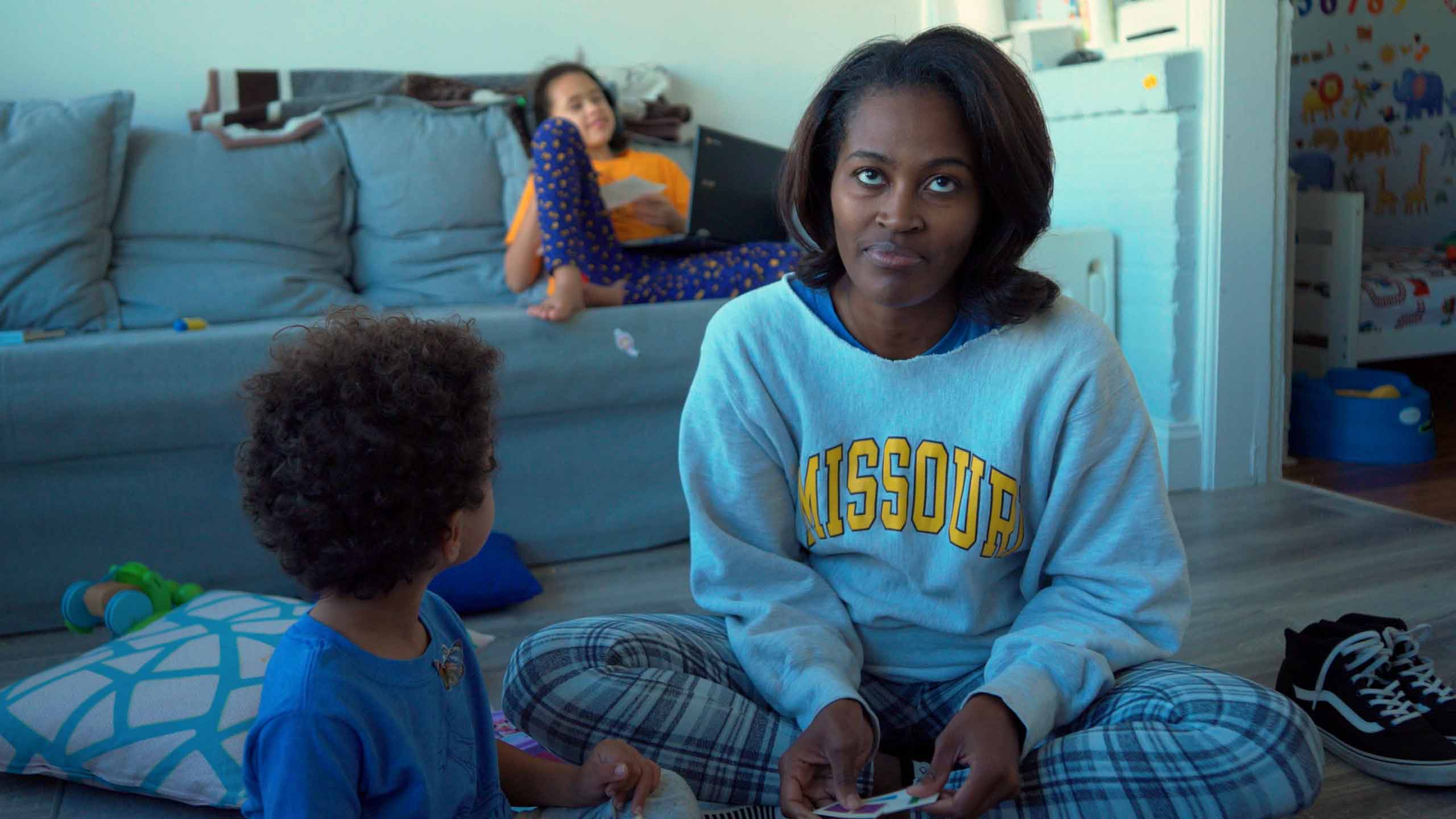 A Black woman looks behind the camera lens as she sits on the floor, working to parent two children.